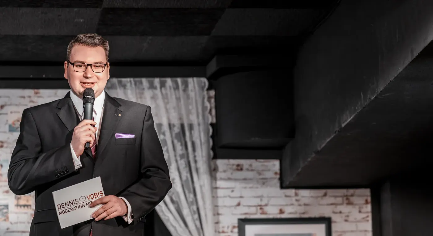 Dennis Vobis moderiert im Anzug mit Mikrofon vor Publikum